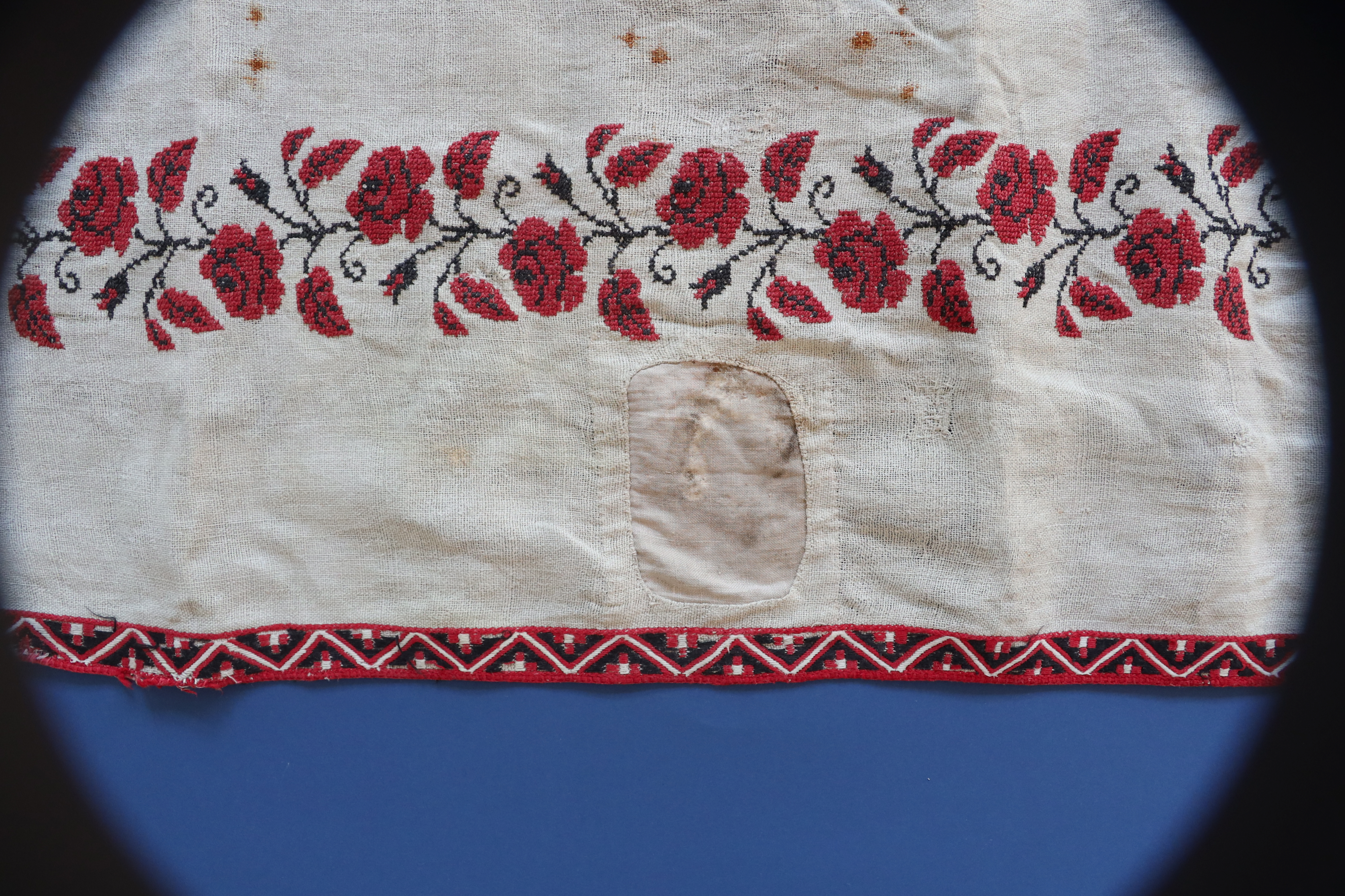 Trzy, stopniowe ujęcia na tzw. bieżnik – noszący ślady użytkowania i wielokrotnych napraw podłużny obrusik. Sfotografowany na arkuszu granatowego tła nie mieści się nim, wychodząc na rozwinięte pod nim tło białe. Zbliżenia pokazują zarówno floralny wzorzec dookoła jak i ślady szeregu ​ cerowań ale też zalań i przypaleń.