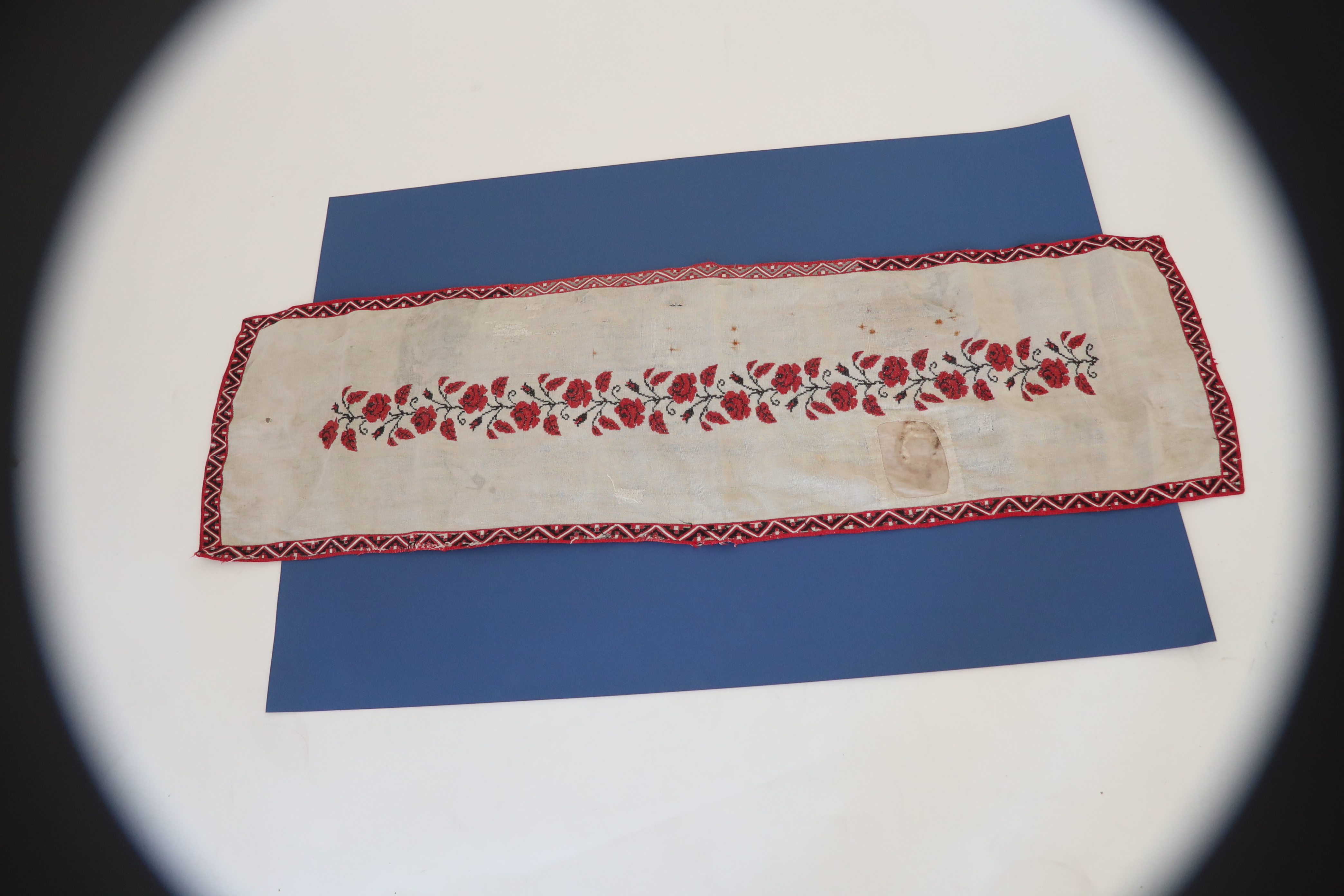 Trzy, stopniowe ujęcia na tzw. bieżnik – noszący ślady użytkowania i wielokrotnych napraw podłużny obrusik. Sfotografowany na arkuszu granatowego tła nie mieści się nim, wychodząc na rozwinięte pod nim tło białe. Zbliżenia pokazują zarówno floralny wzorzec dookoła jak i ślady szeregu ​ cerowań ale też zalań i przypaleń.