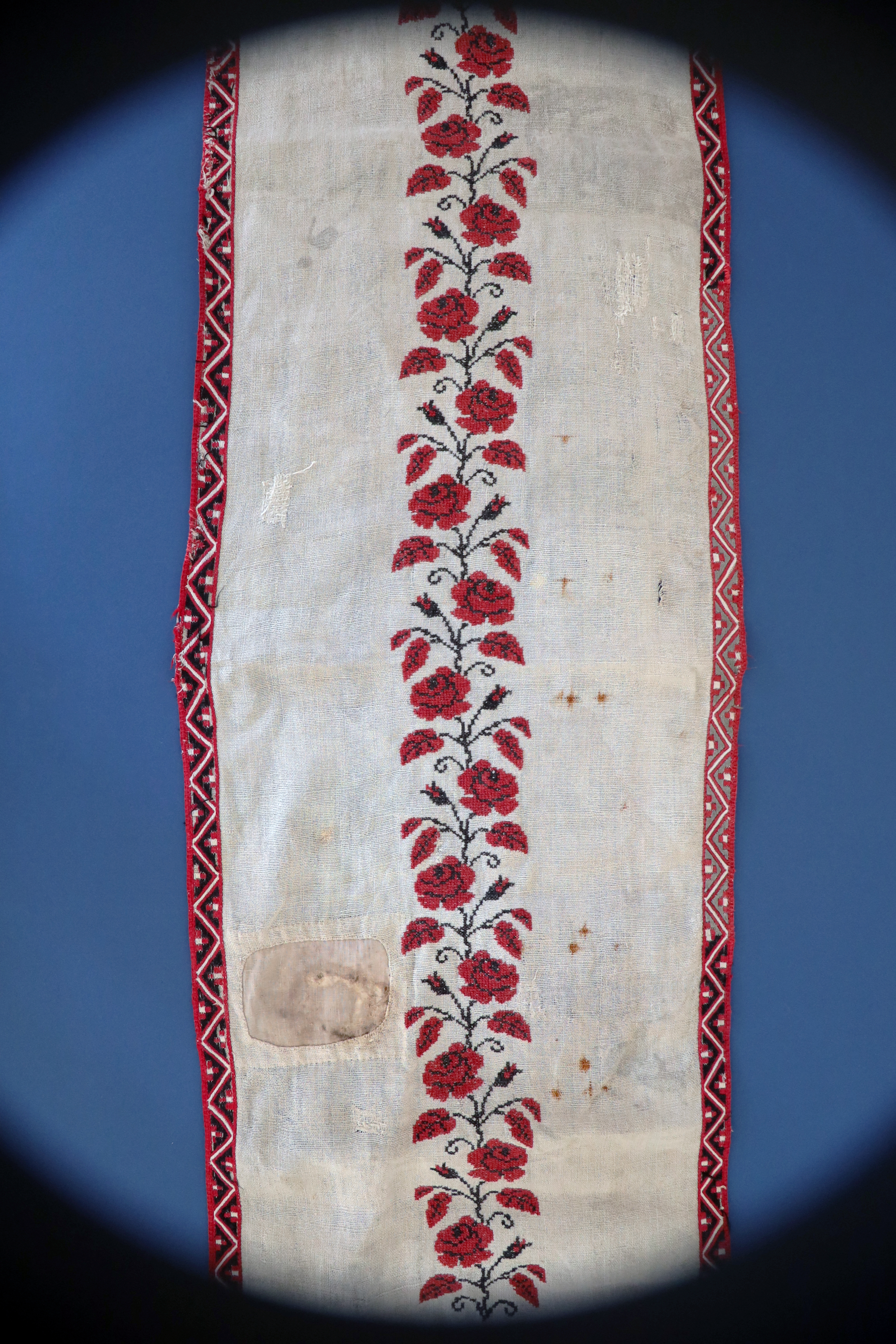 Trzy, stopniowe ujęcia na tzw. bieżnik – noszący ślady użytkowania i wielokrotnych napraw podłużny obrusik. Sfotografowany na arkuszu granatowego tła nie mieści się nim, wychodząc na rozwinięte pod nim tło białe. Zbliżenia pokazują zarówno floralny wzorzec dookoła jak i ślady szeregu ​ cerowań ale też zalań i przypaleń.